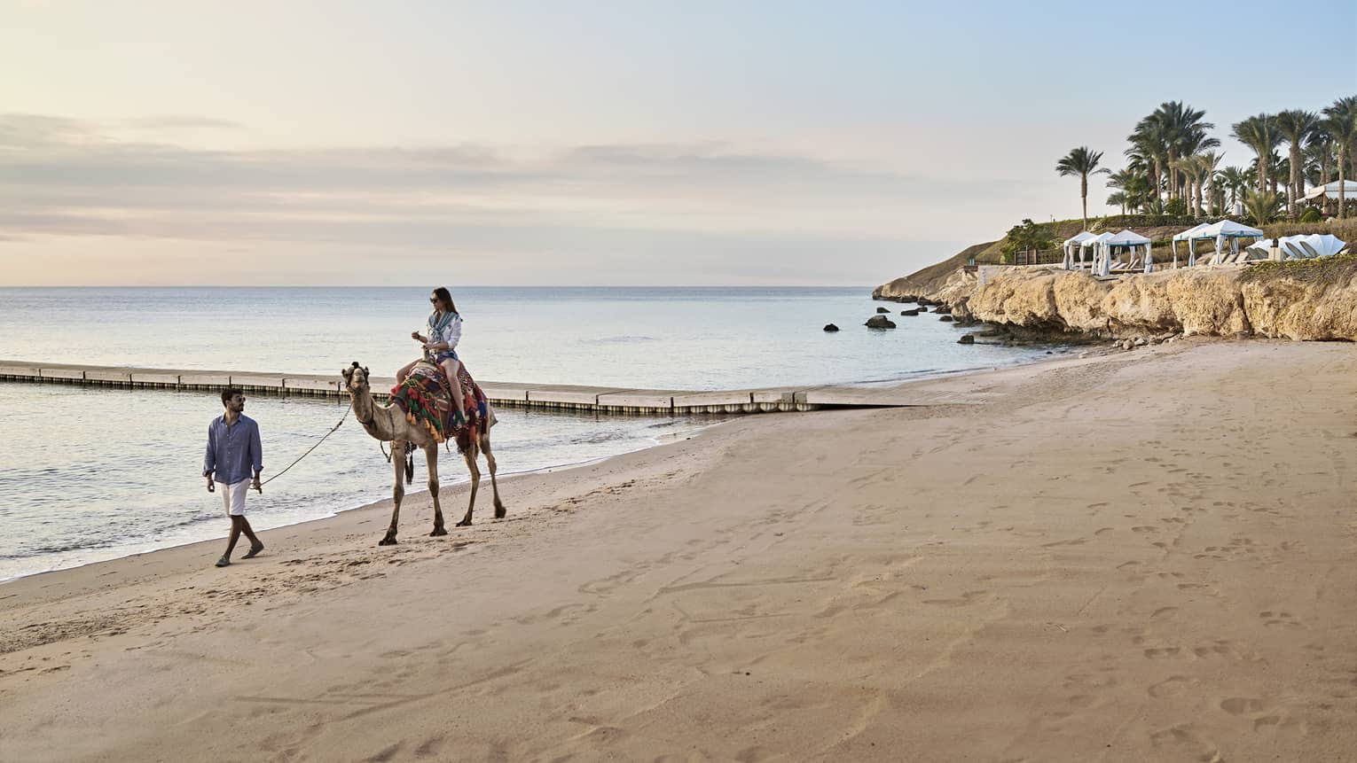 A woman rides a camel, led by a man, along the beach at Four Seasons Sharm El Sheikh
