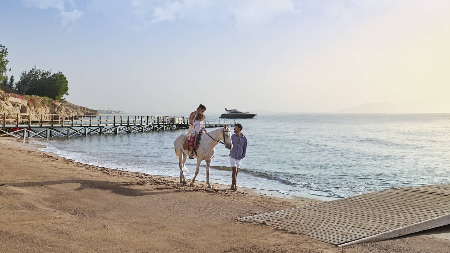 An adult and child on a white horse on a beach, led by another guest, with a wooden pier and a yacht visible in the distance.