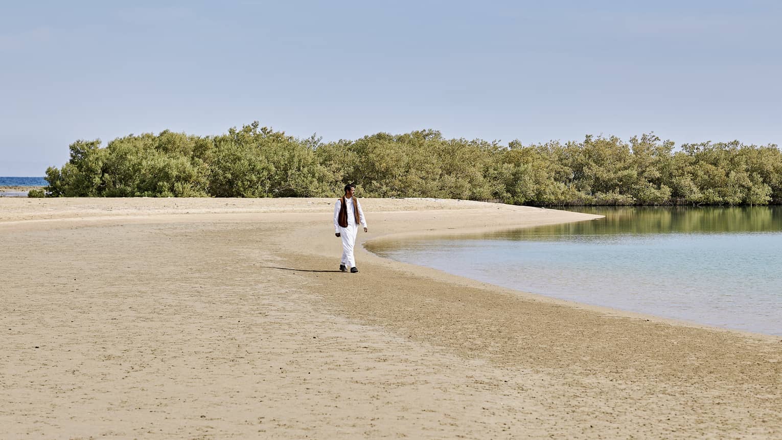 A guest wearing loose white clothing looks out across rippling water as they walk along a curving sandy shoreline with trees.