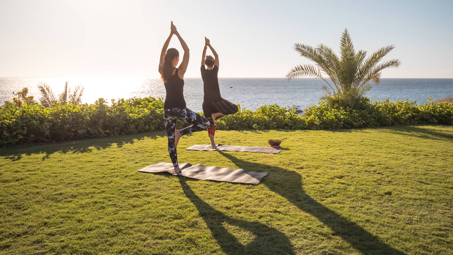 Two people enjoy yoga outdoors by the sea with the sun rising in the sky at a luxury hotel