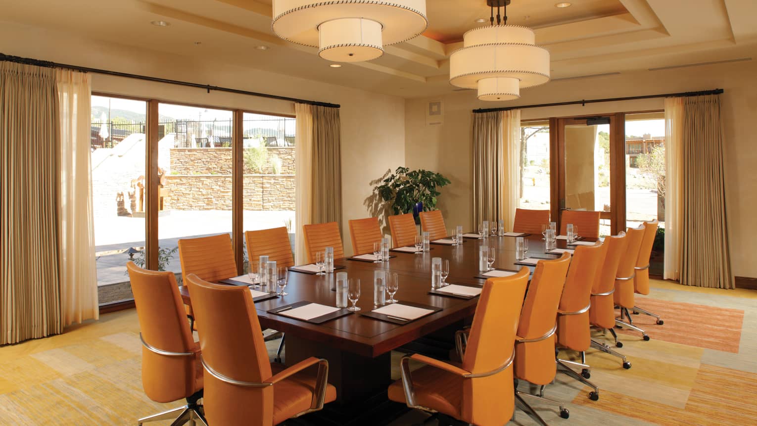 Executive board table surrounded by orange chairs, white ceiling lights, floor-to-ceiling windows overlooking terrace 