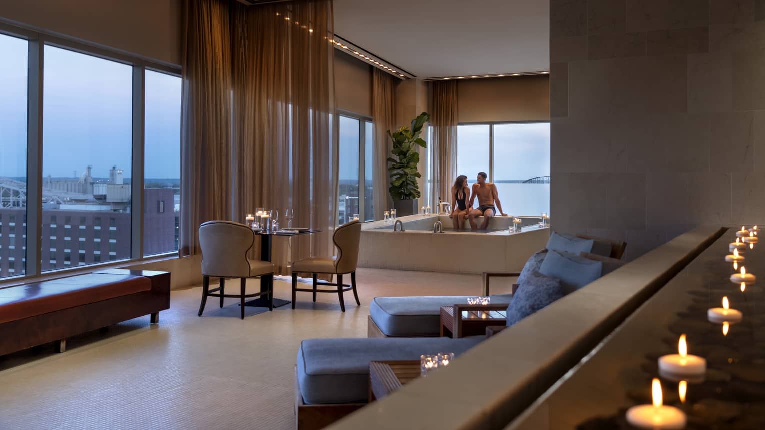 A couple enjoying a candlelit hot tub in a luxury hotel spa, with elegant seating, soft lighting and large windows offering a view of a distant bridge