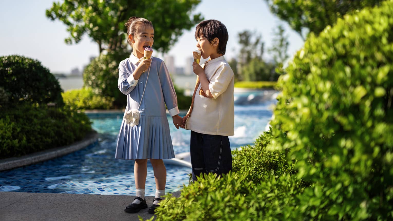 Young boy and girl hold hands by outdoor pool, eating ice cream, at luxury hotel