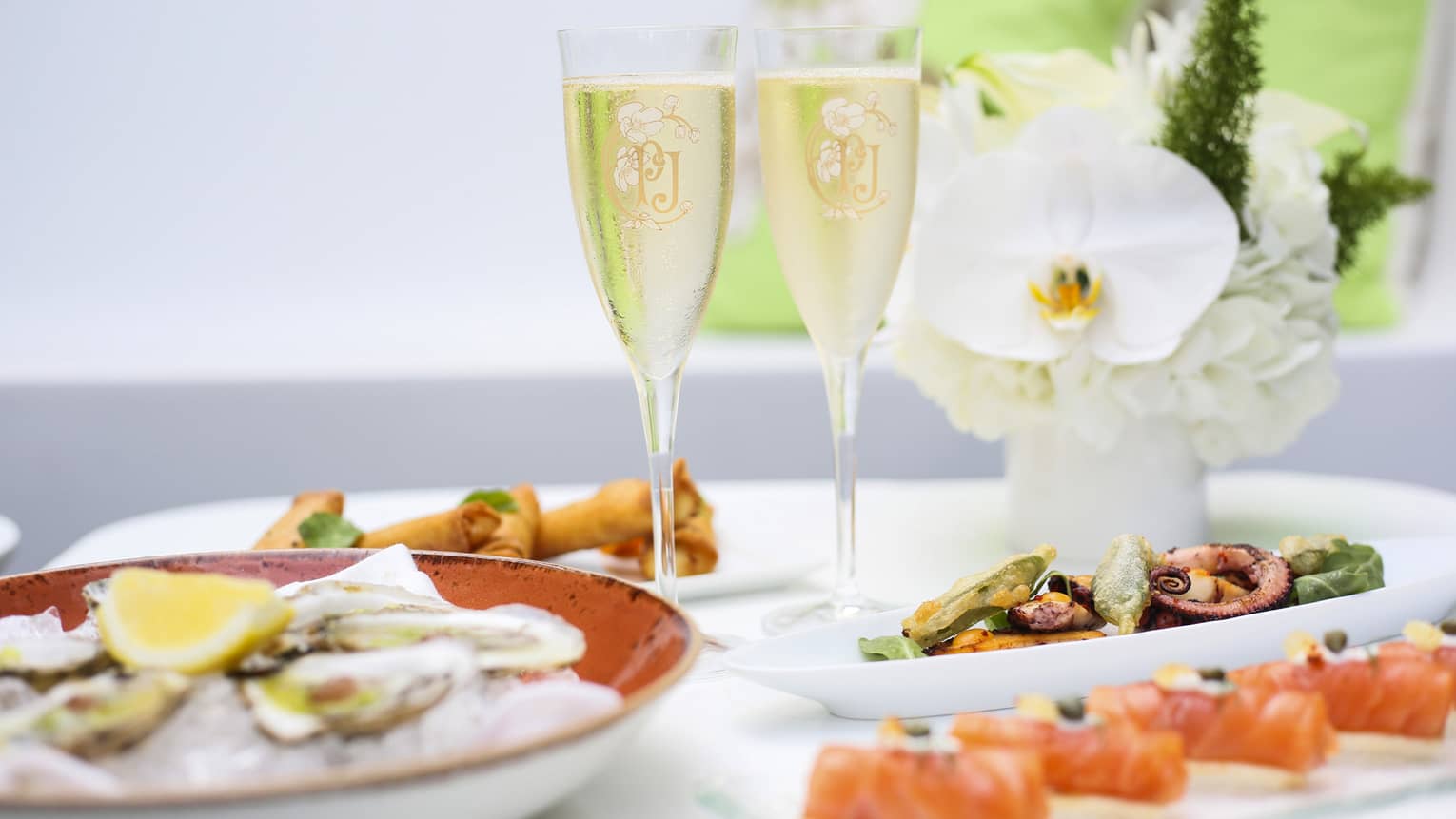 Two glasses of Champagne, flowers, trays with oysters and canapes on white table