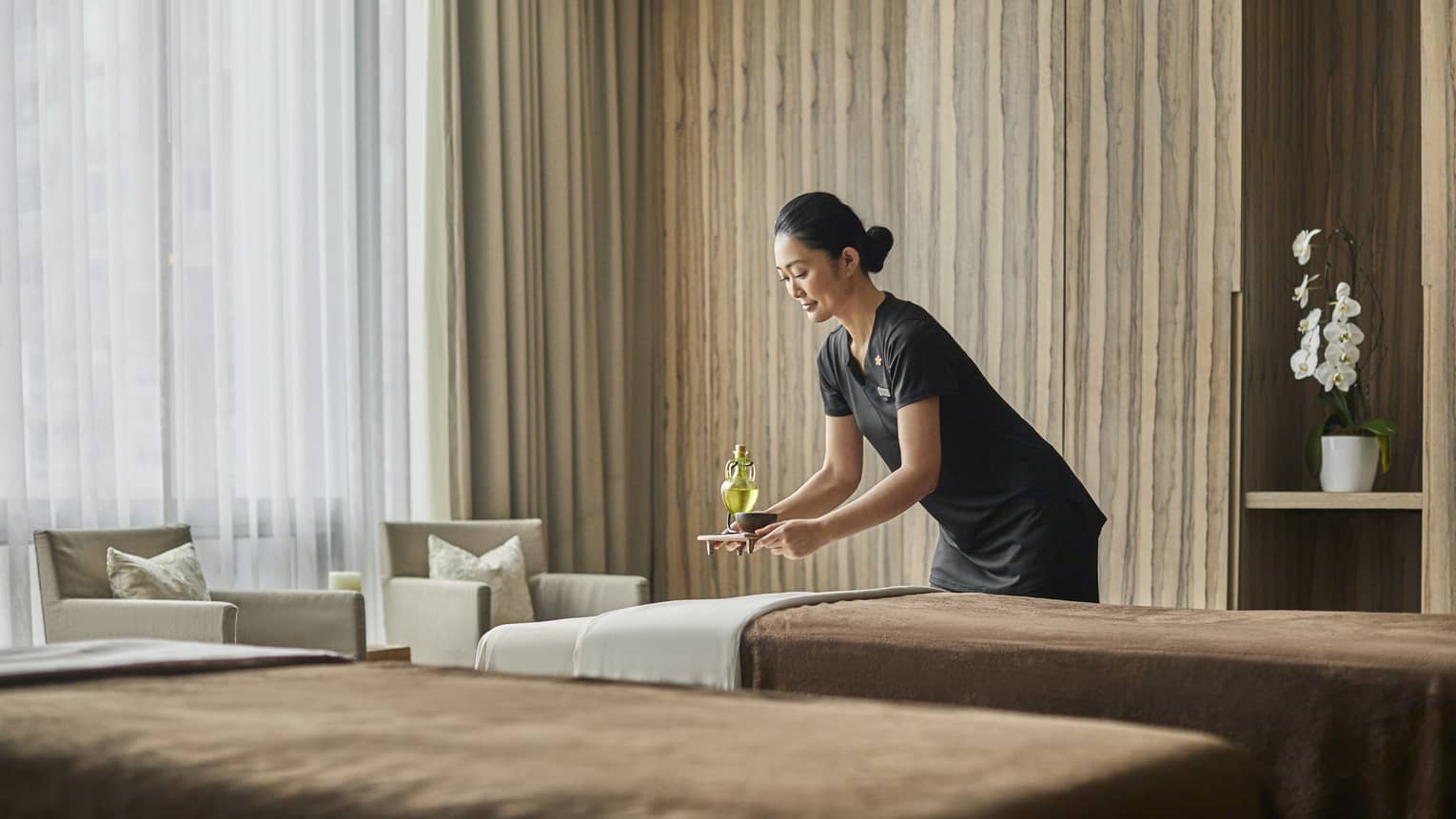 A female masseuse brings a drink to sit behind one of two massage beds. 