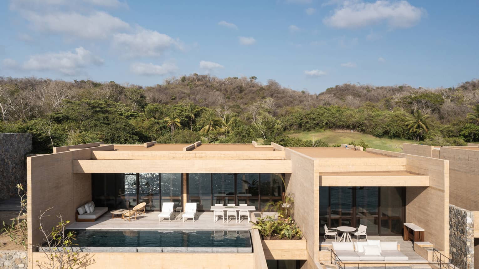 A modern beachfront villa with minimalist architecture, featuring a private pool, sun loungers and outdoor seating areas. The villa is surrounded by lush greenery and hills under a clear sky.