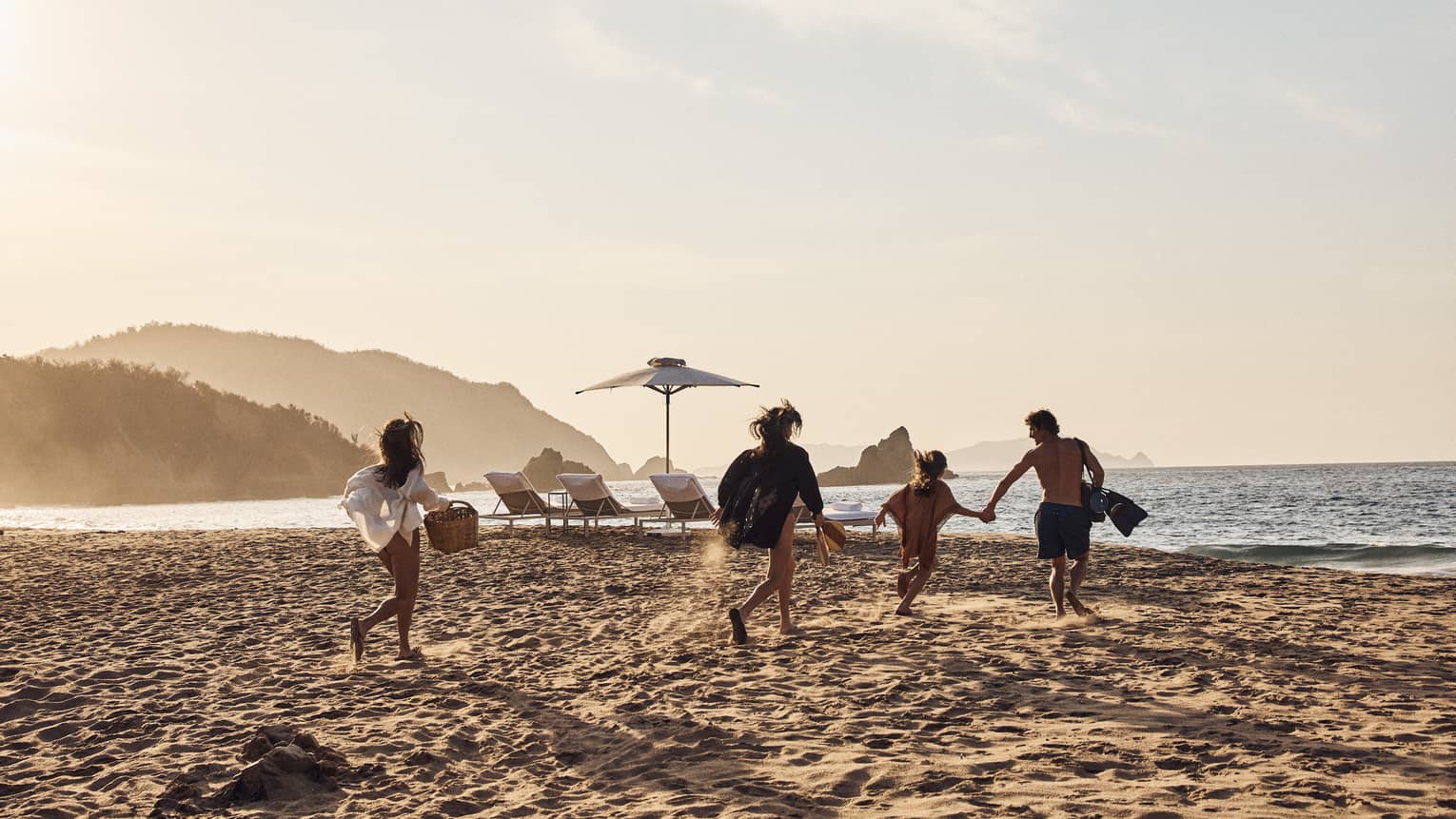 A group carrying various beach supplies runs across the sand toward the water, empty lounge chairs prepared for them. 