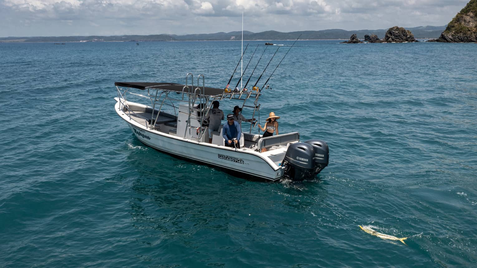 Guest reels in a large yellow fish from the back of a boat in open water near rocky islands, while two friends look on.