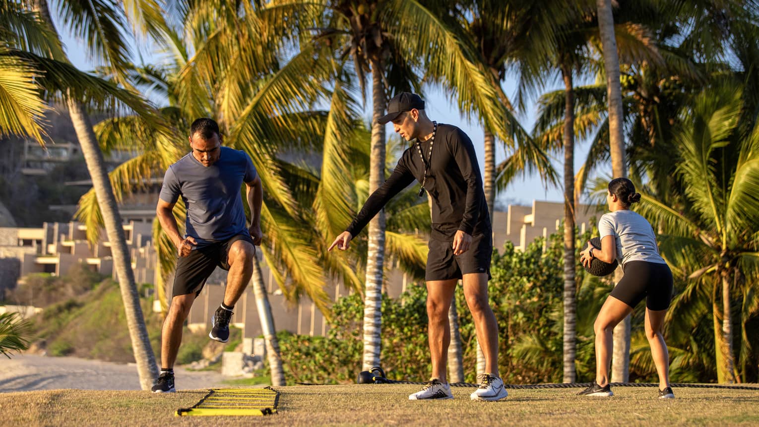 Fitness instructor speaking to two guests while they exercise outside surrounded by palm trees.