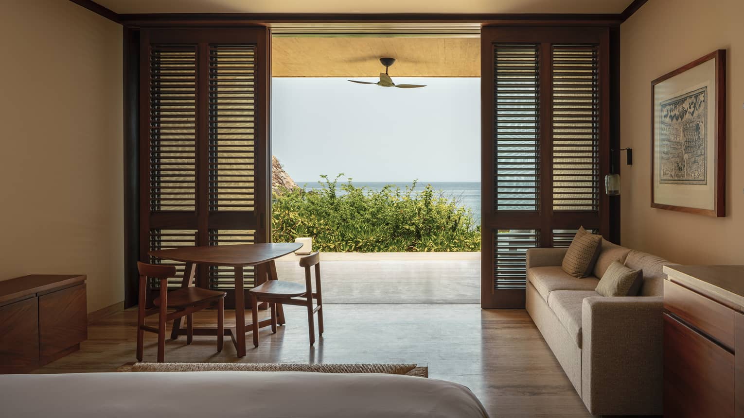 Cozy bedroom with wooden shutters, a sofa and a small dining table. The room opens to a private terrace with ocean views, framed by greenery and a ceiling fan.