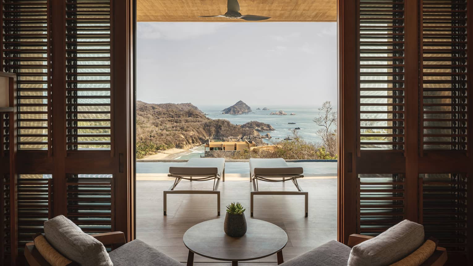 A tranquil ocean view from a luxury villa, featuring two lounge chairs beside an infinity pool, framed by wooden shutters, overlooking rocky islands and a sandy beach