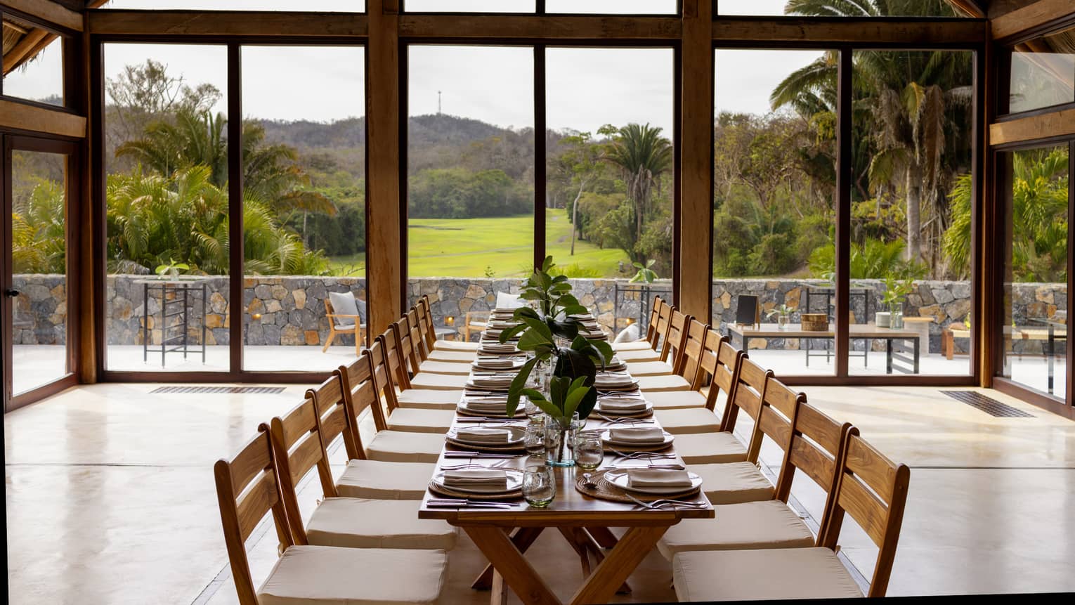 Room with floor-to-ceiling windows and a view of greenery and the mountains, with a rectangular table and place settings.
