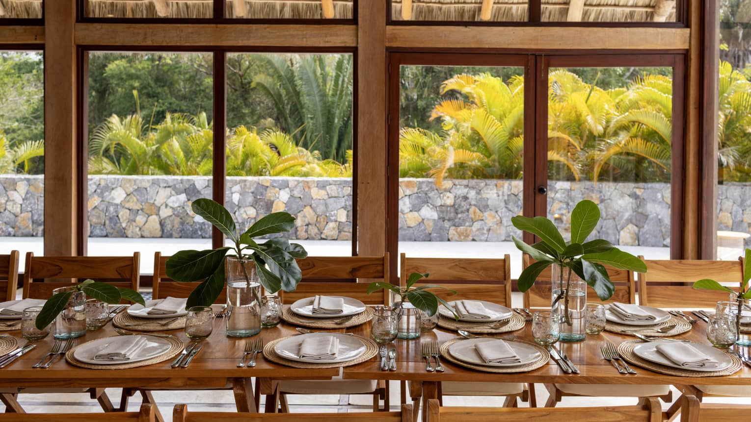 Wooden table and chairs with plates and glasses at each seat and plants lining the table.