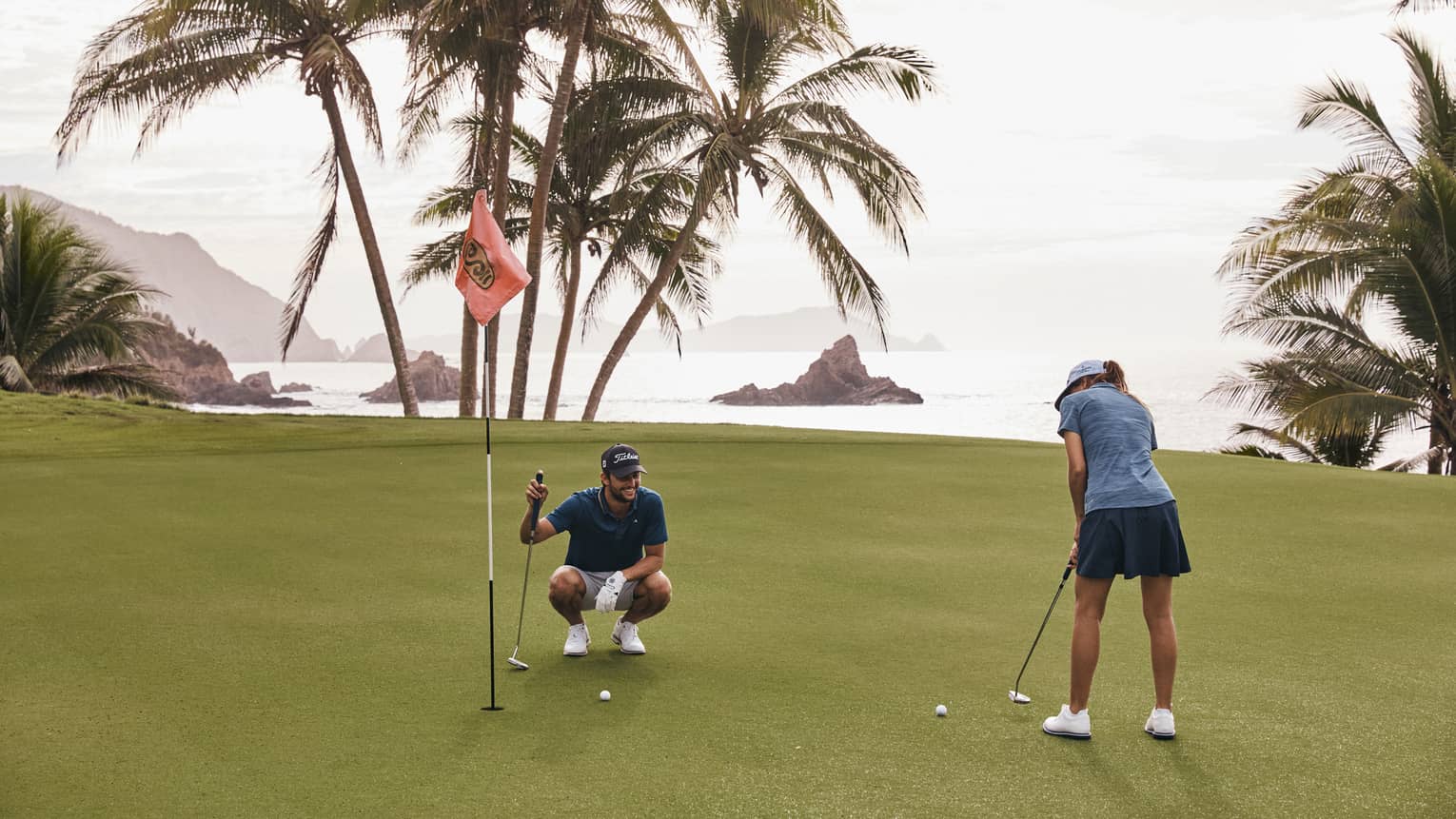 On a golf green surrounded by water and palm trees, one golfer putts while another kneels beside the hole and watches.