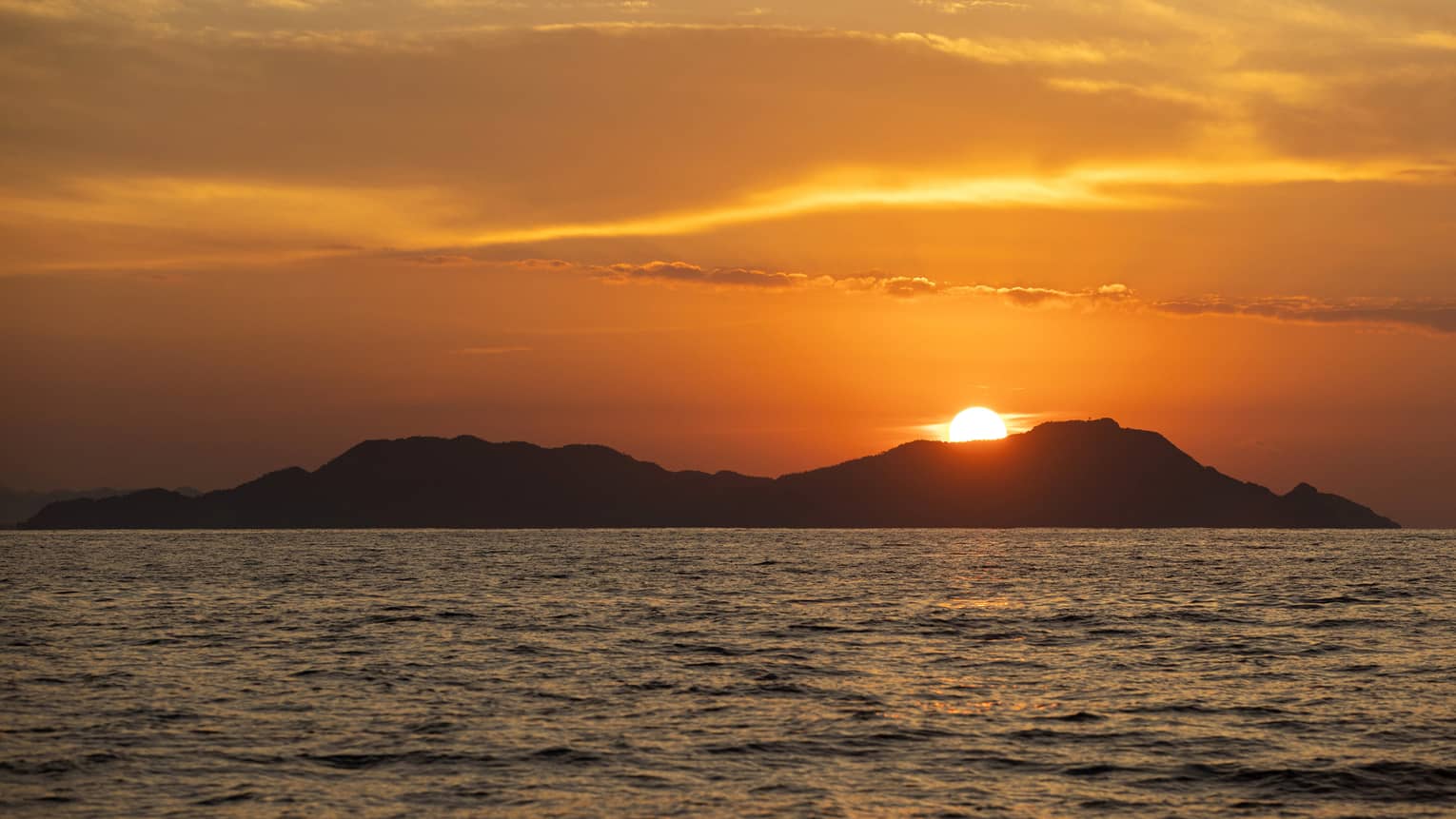 Beyond gently rippling waves, the blazing sun sets behind a hilly island, wispy clouds in the sky awash in its orange glow.
