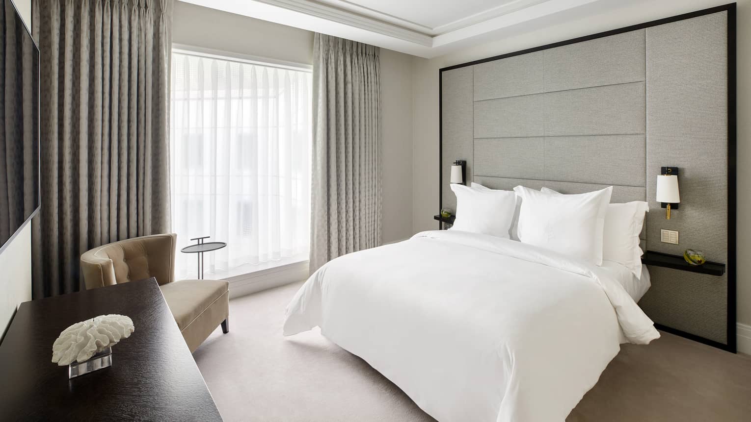 A bed with white sheets, large window chair and counter in a room.