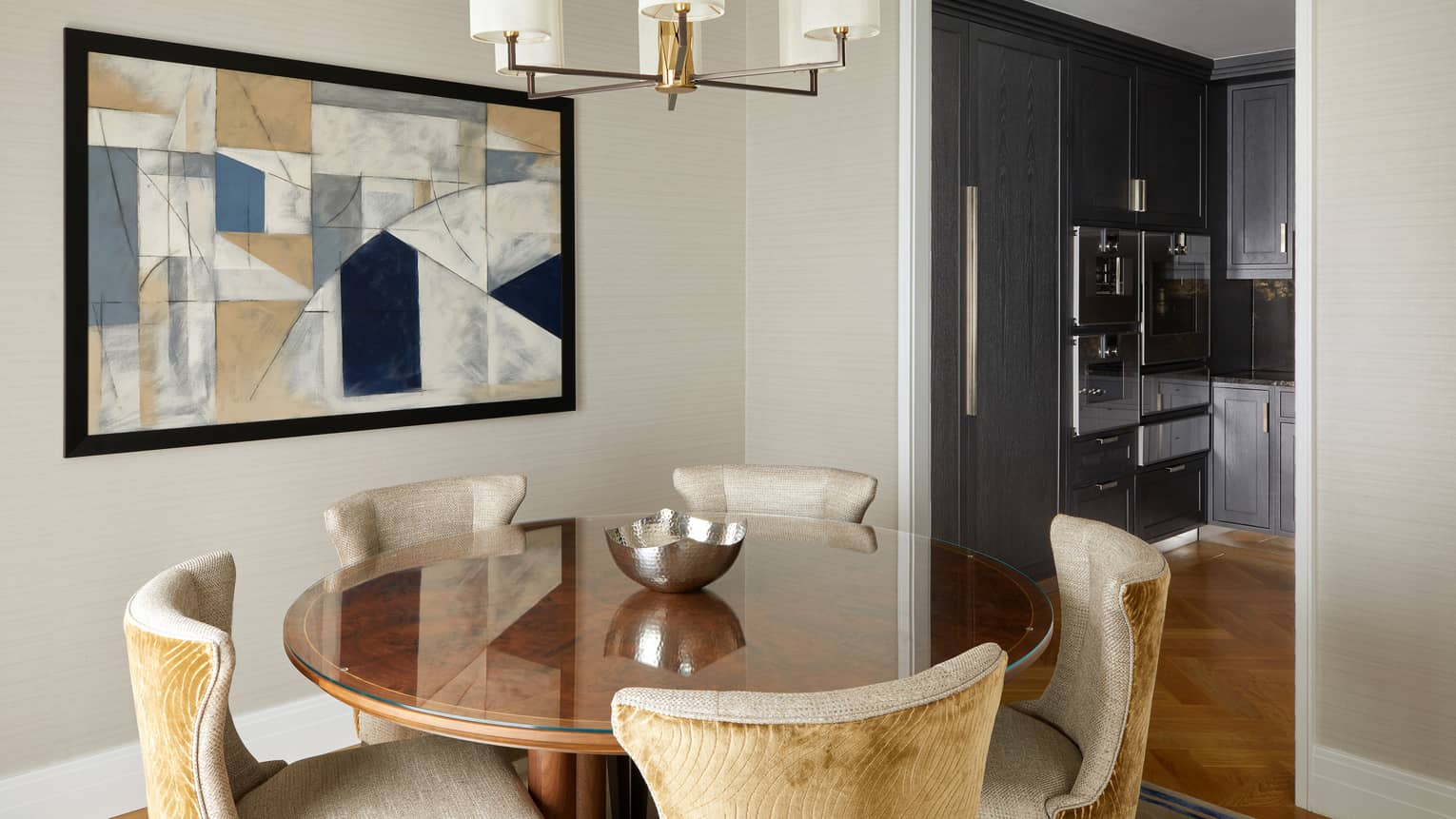 A round table with five chairs below a hanging light and near a kitchen.