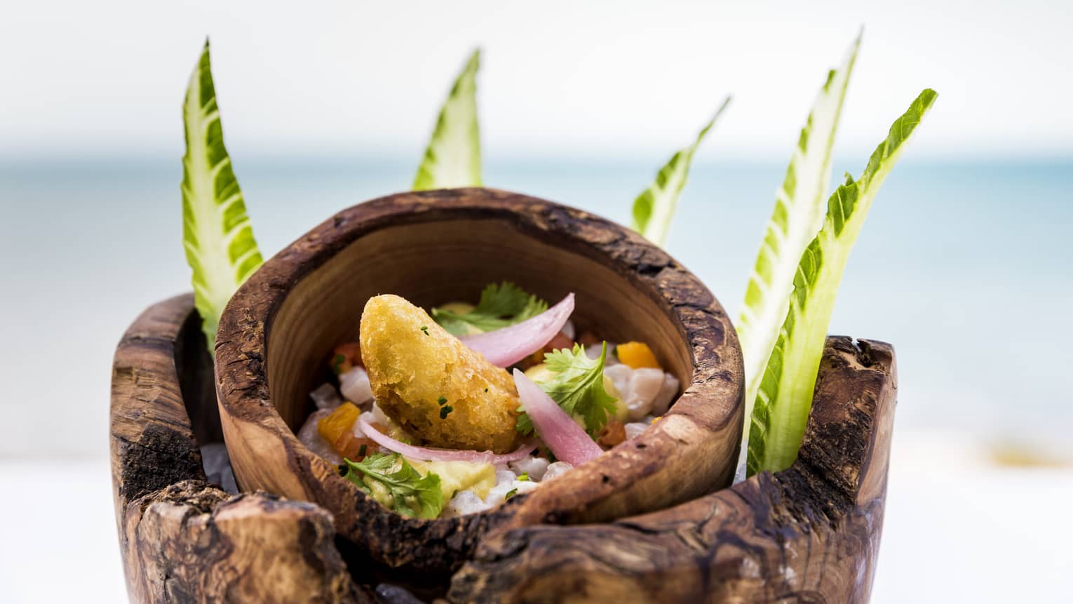 Cuisine served in wooden bowl, surrounded by lettuce leafs with ocean in background 