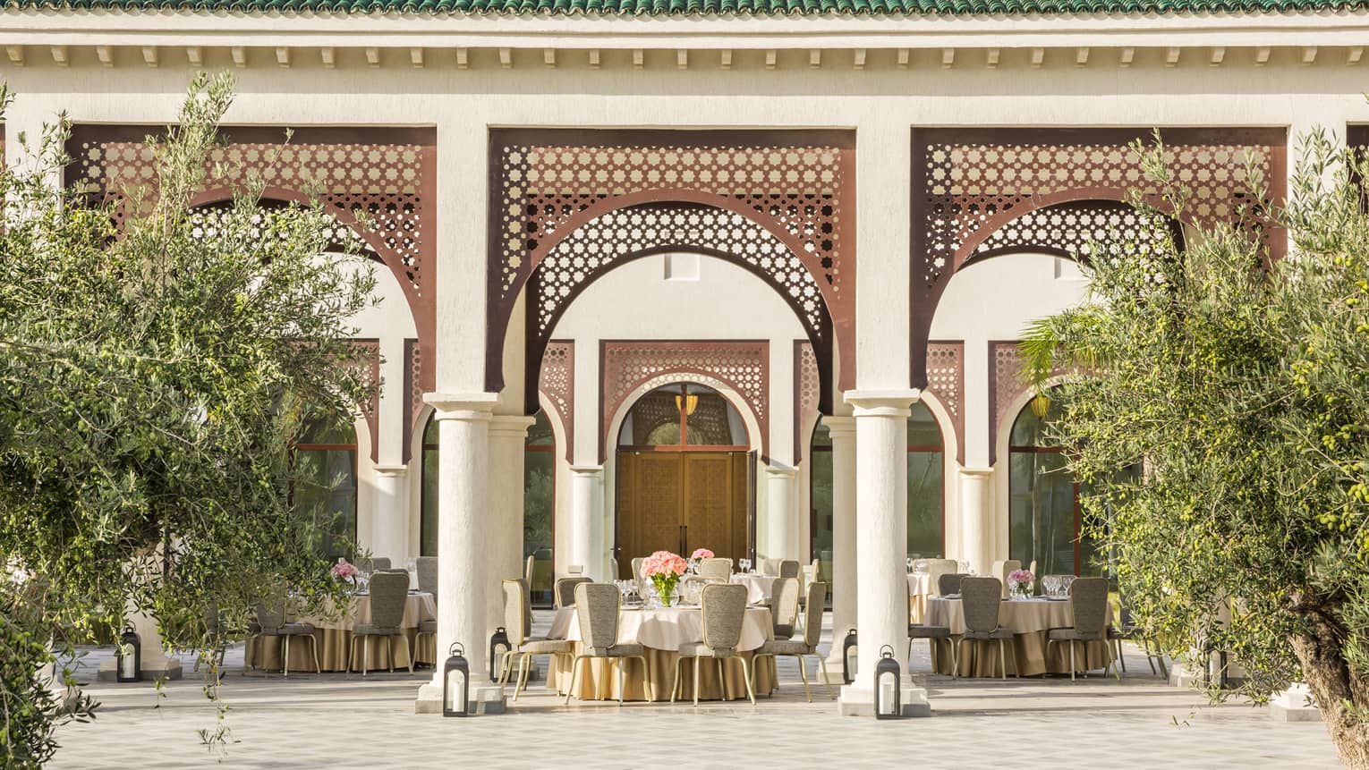 Round tables and chairs with vases of flowers set on covered patio with trees, lanterns 