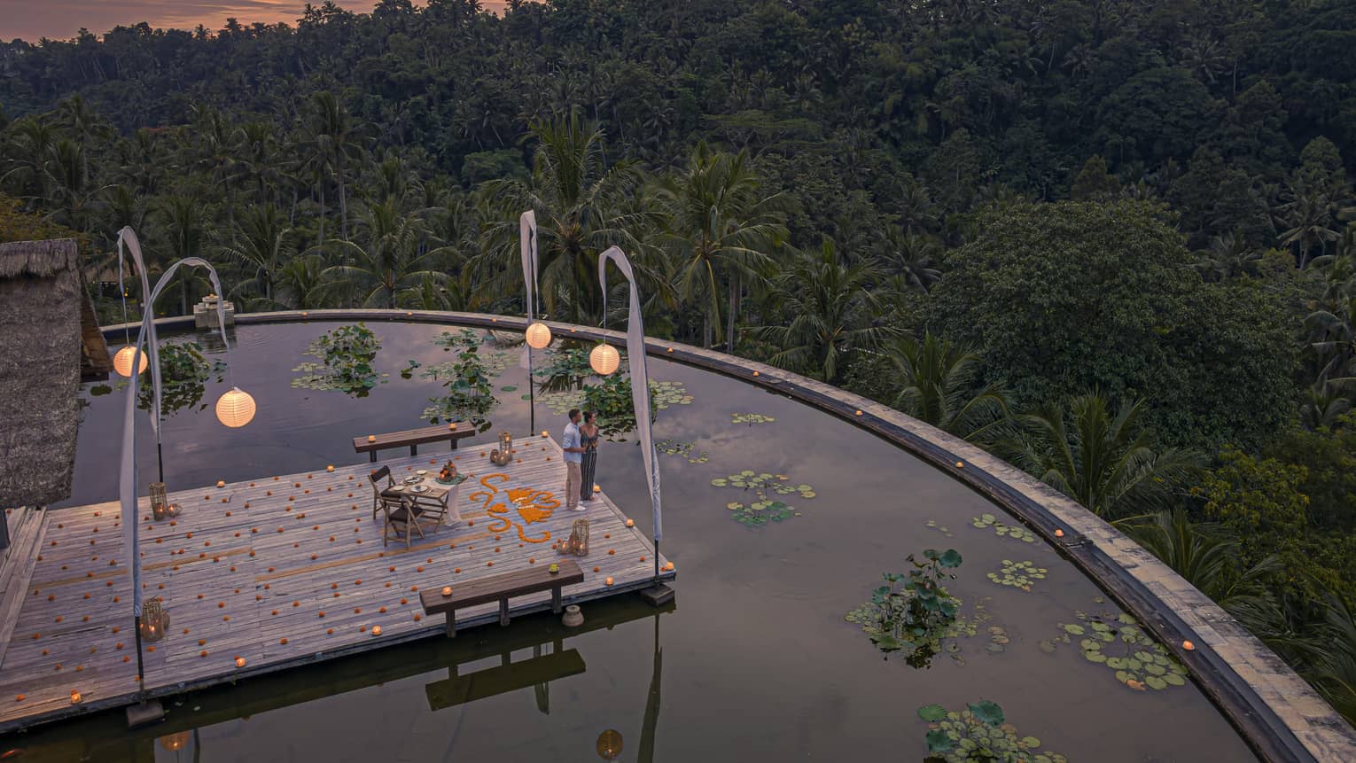 Couple stands on dock at the infinity edge lotus pond overlooking dense forest at dusk
