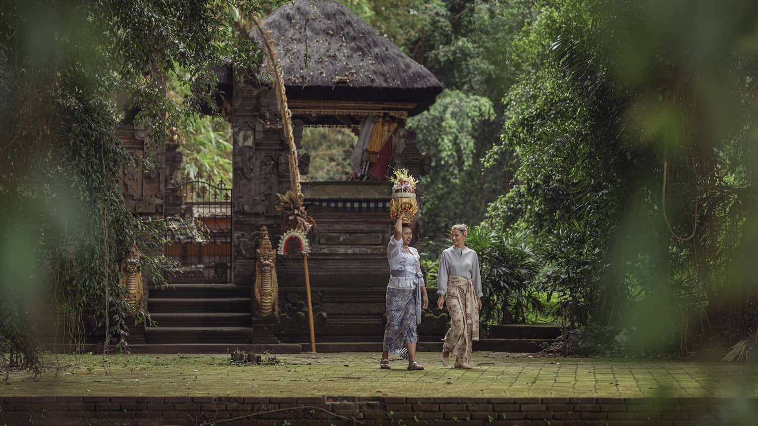 Two women wearing batik skirts walk by a temple in a lush tropical area as one of them carries a golden tray on her head.