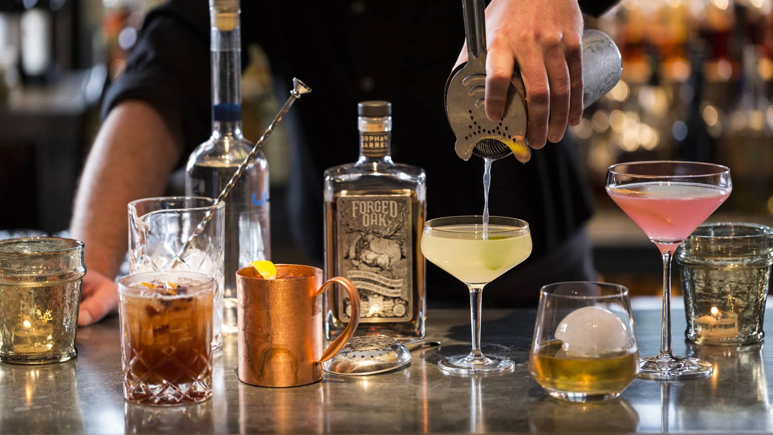 Various cocktail glasses and liquor bottles on bar as mixologist pours martini out of shaker