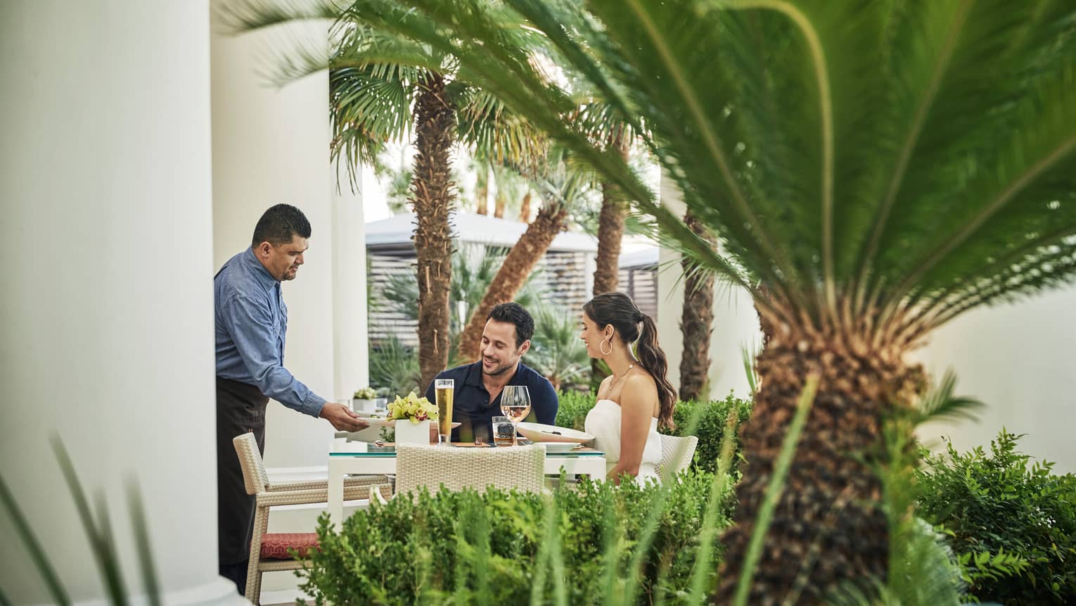 A man serves a man and woman sitting at outdoor table, surrounded by palm trees