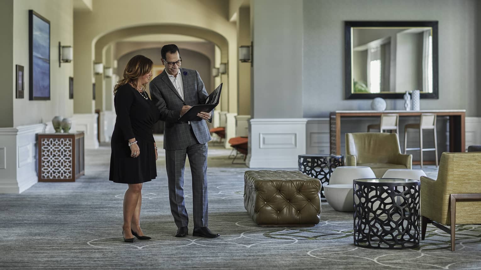 A man and woman dressed in business attire discuss while looking at a folder of documents in the lobby 