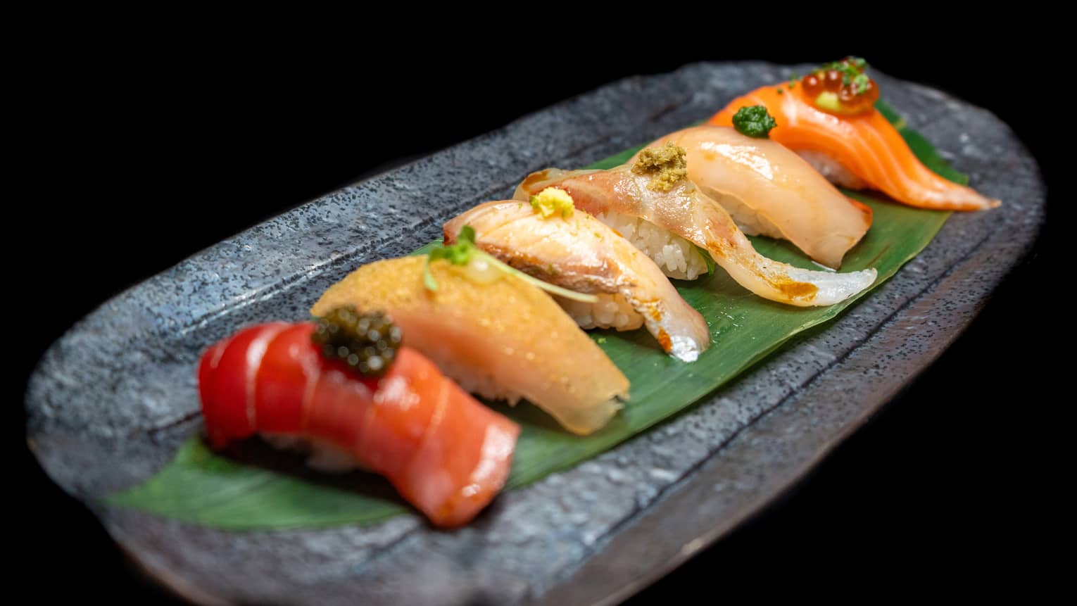 An oblong gray plate holds six pieces of brightly coloured nigiri, set on a long green leaf.