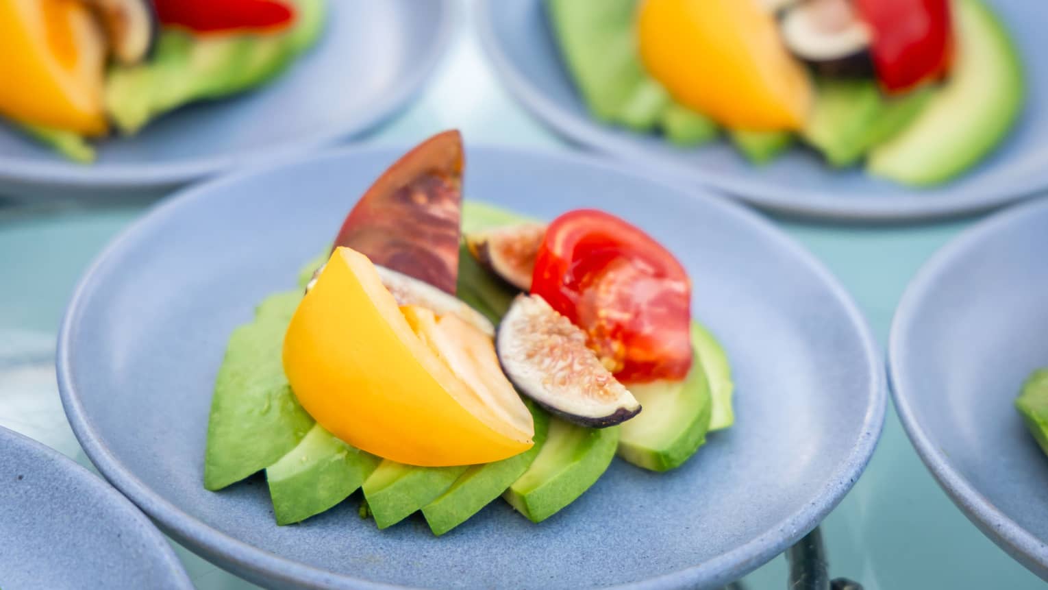 Three small light blue plates each with slices of avocado, multi-coloured tomato and figs