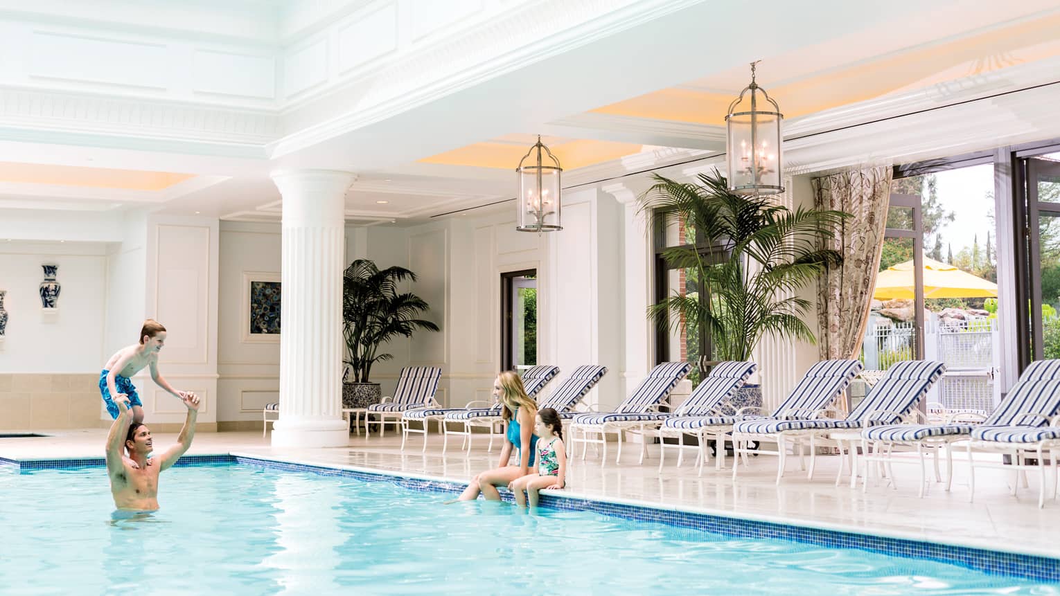 Family swimming in indoor pool, mom and daughter on ledge, boy on dad's shoulders
