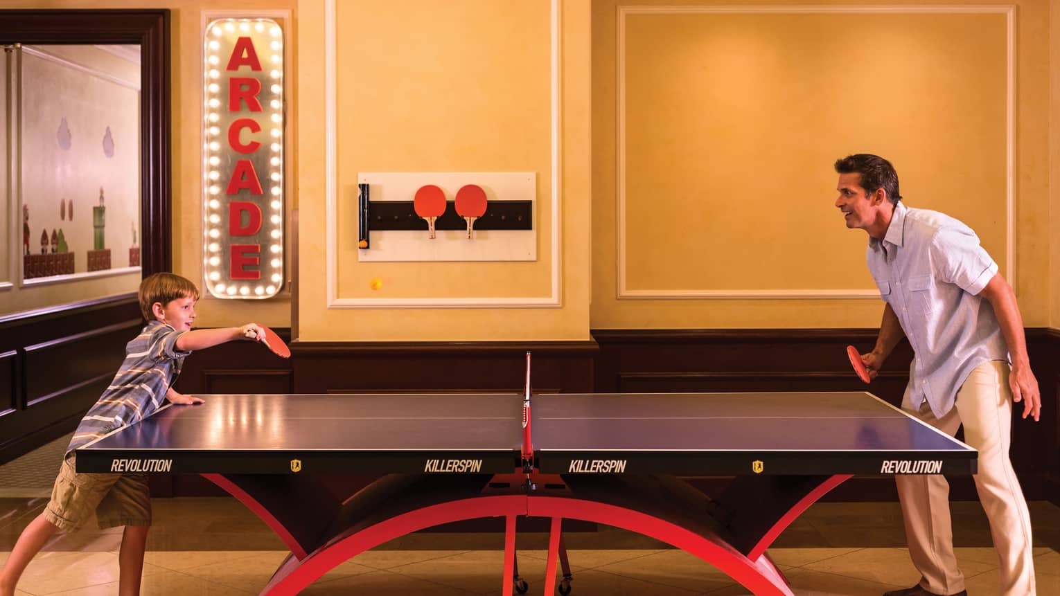 Man, young boy play table tennis under arcade sign with lights in Game Room