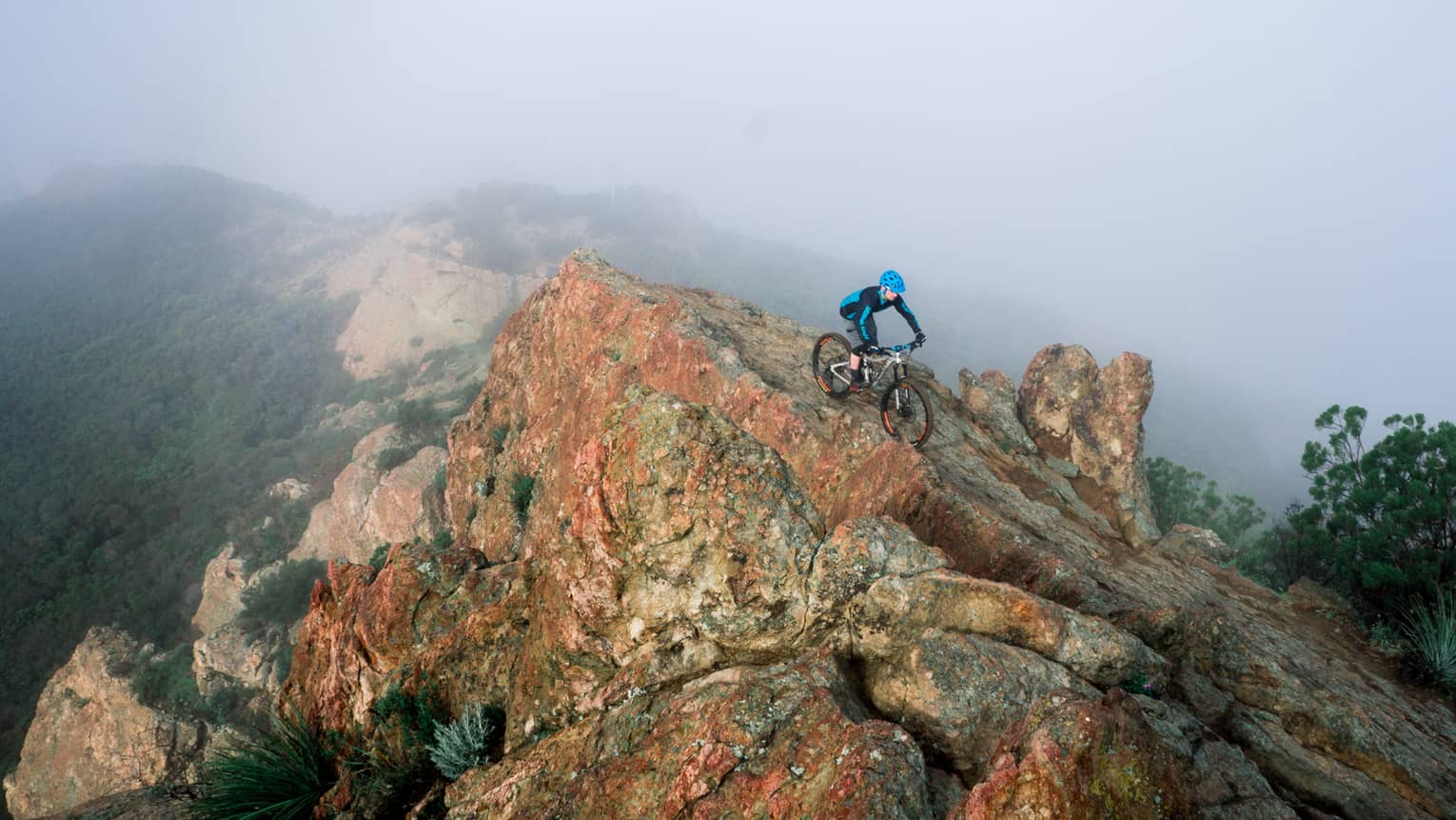 Cyclist rides down large boulder at top of misty mountain