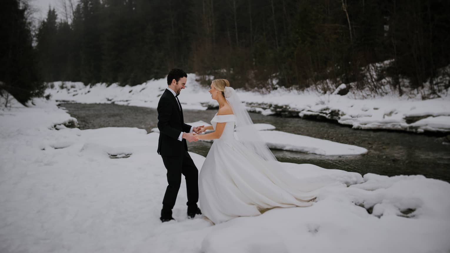 Laughing couple in wedding attire hold hands along the banks of a snowy river