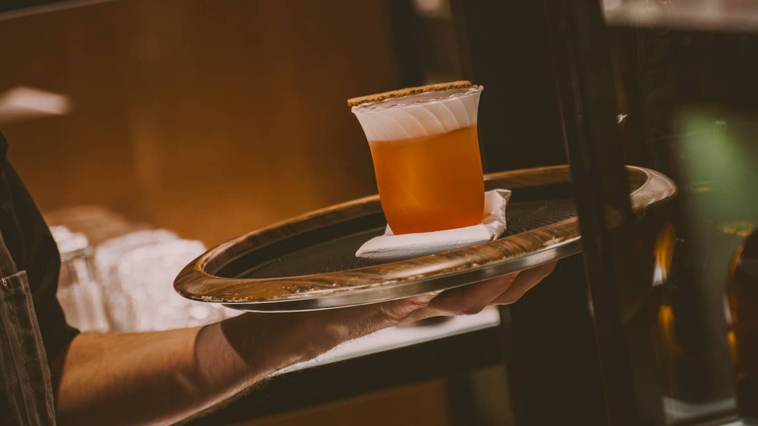 Orange-coloured cocktail served in a scallopped-edge glass being carried on a round silver tray by a server