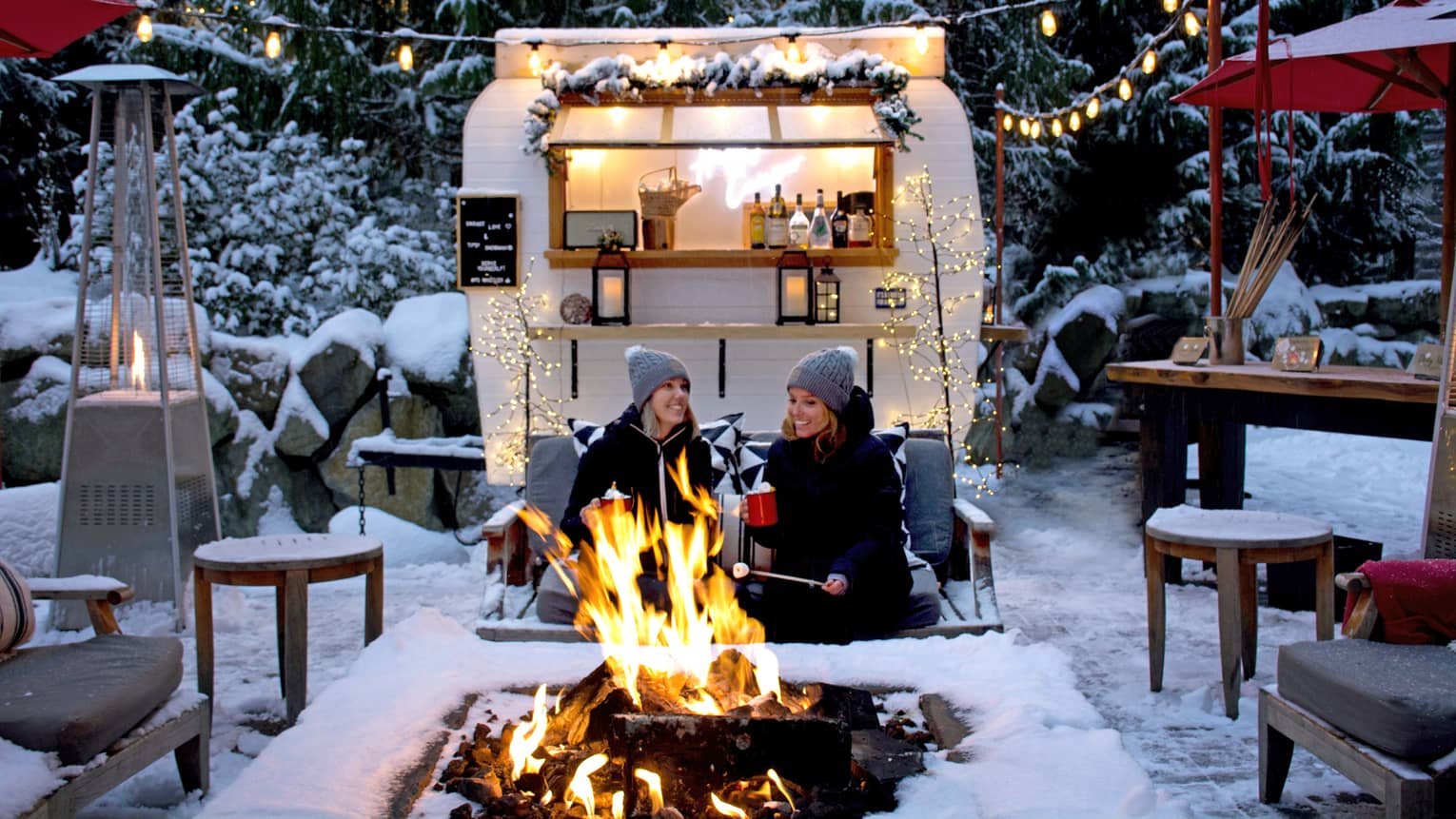 Two women roasting marshmallows at an outdoor fire pit with camper in background