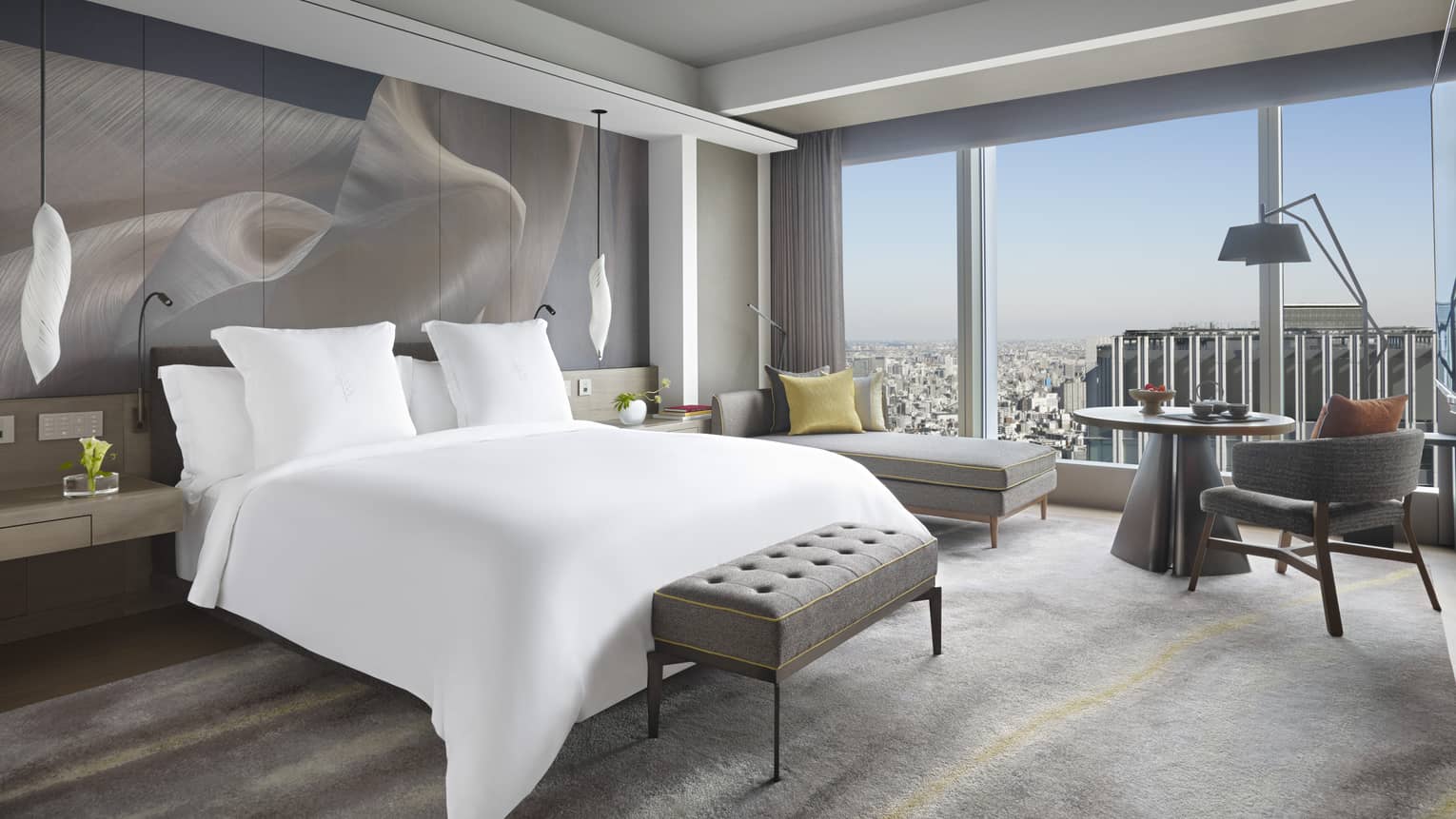 Guest room with king bed, tufted bench, chaise lounge, table with armchair, pendant lights and large window offering a city skyline view