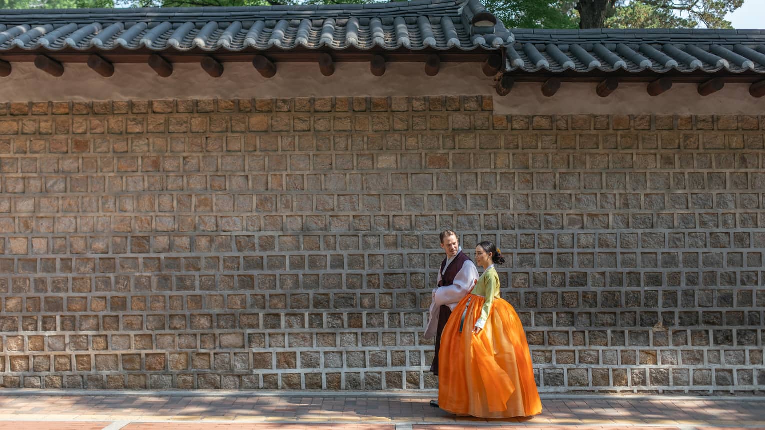 Couple in traditional Korean regalia walk together beside tall brick wall
