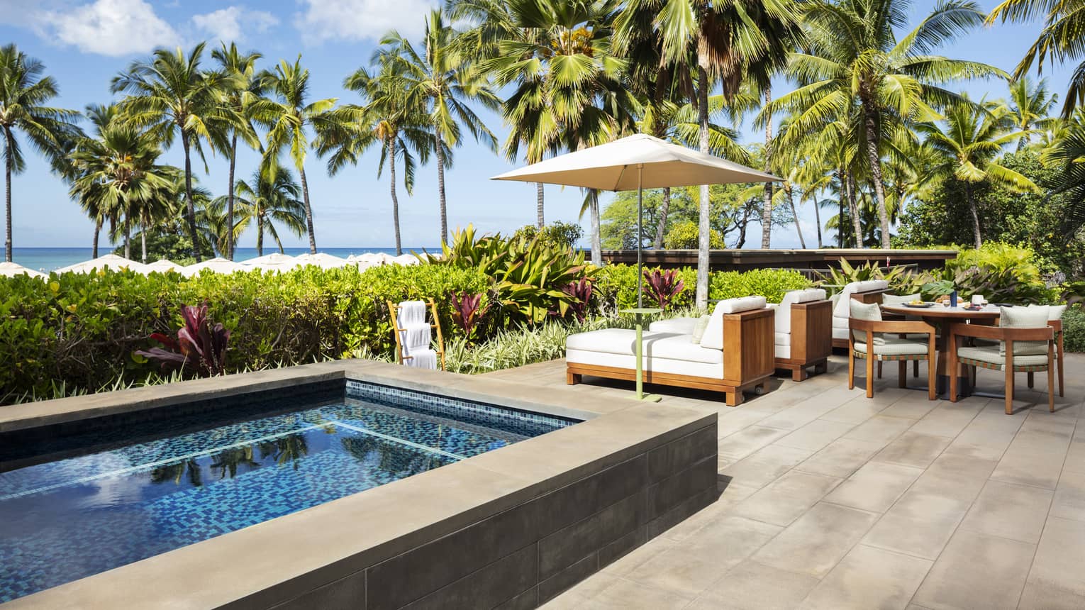 Luxury resort suite private terrace featuring a small rectangular pool, wooden lounge chairs with flush white cushions, and a dining table with palm tree ad ocean views