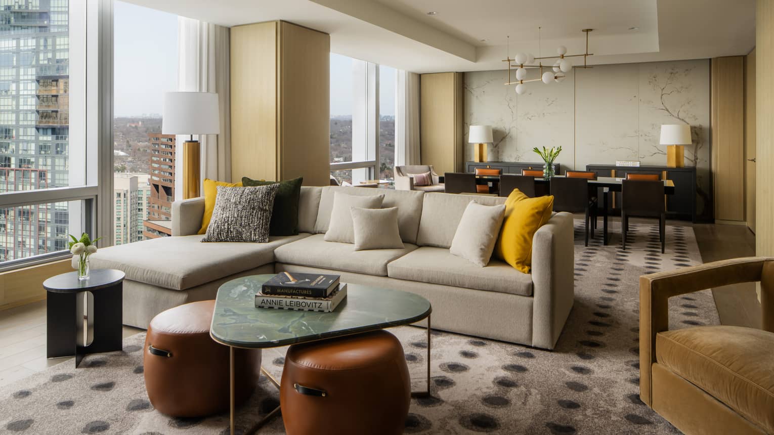 Spacious suite living area with a large sectional sofa, yellow and gray pillows, marble coffee table, and a dining area with elegant décor and city views