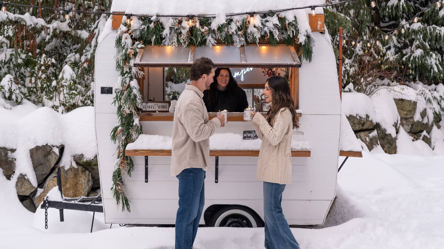 A couple holds mugs of hot chocolate outside of a camper in a snowy courtyard