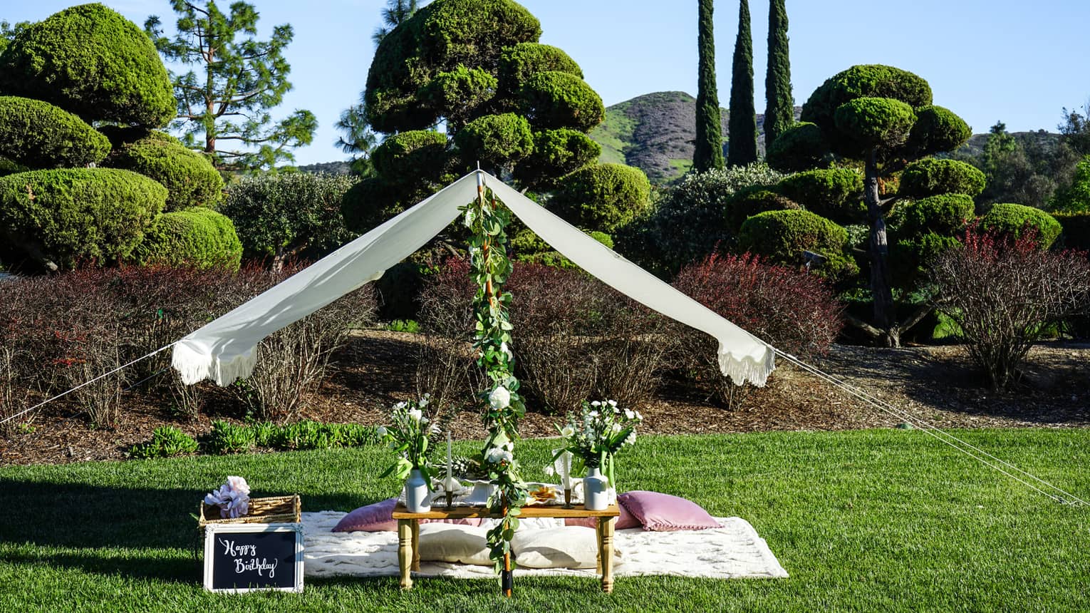 Picnic set up on a manicured lawn with flowers and a white canopy over the blanket