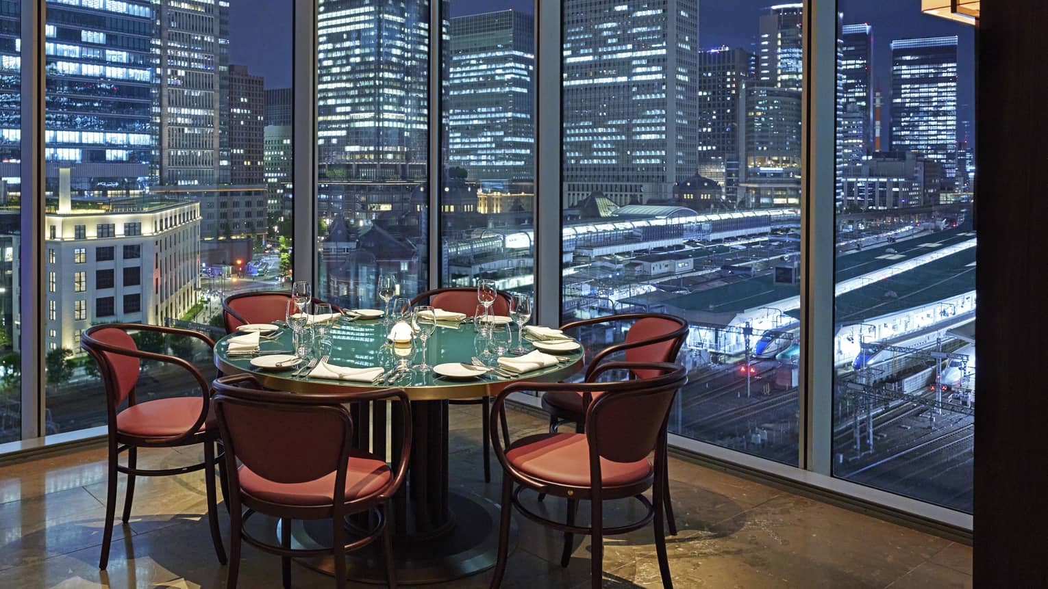 Luxury boutique hotel dining setup with a round table elegantly set for guests, offering a panoramic city view at night through tall glass windows