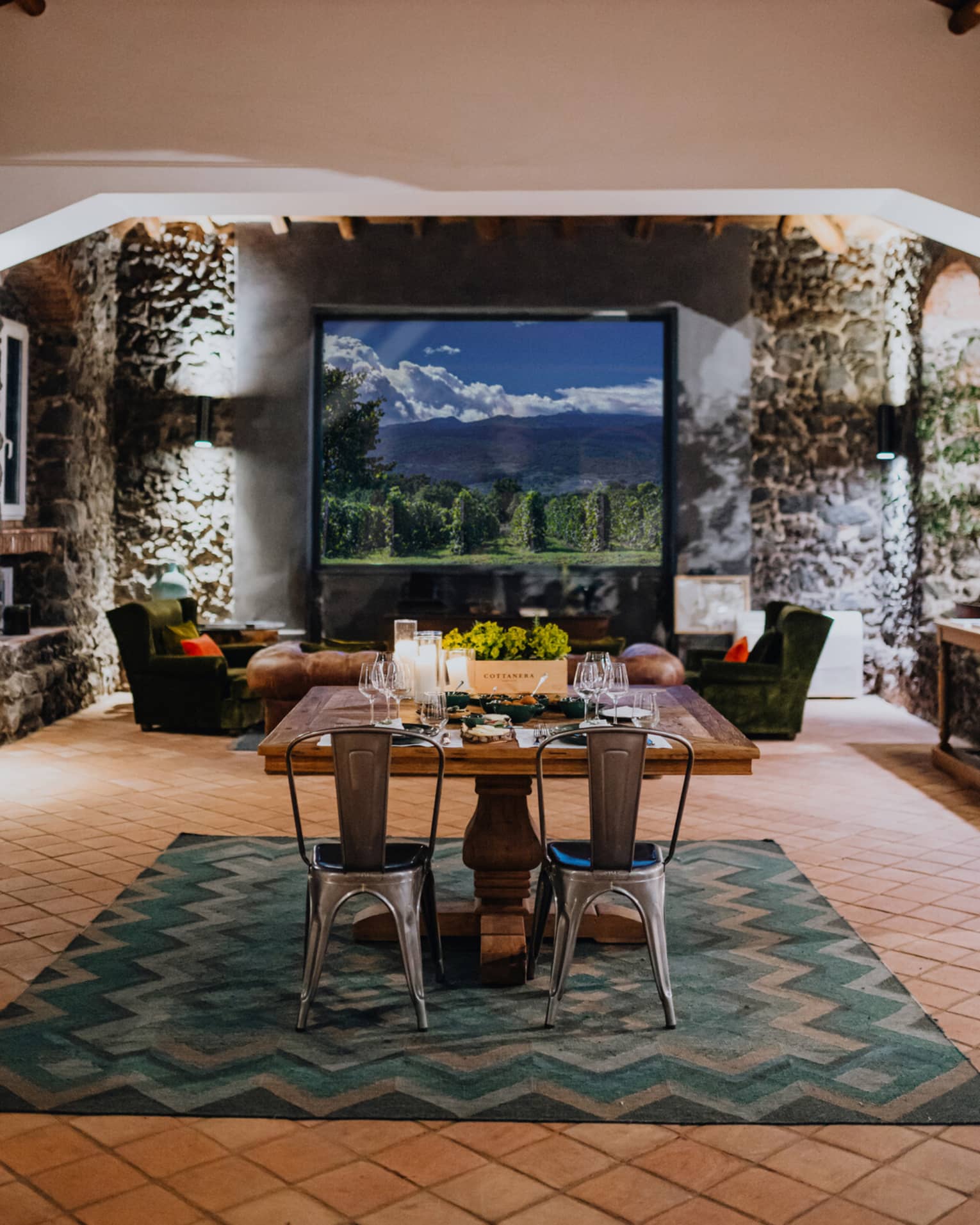 A wooden dining table set with floral arrangements and glassware,  in a room with a window overlooking mountains and a vineyard