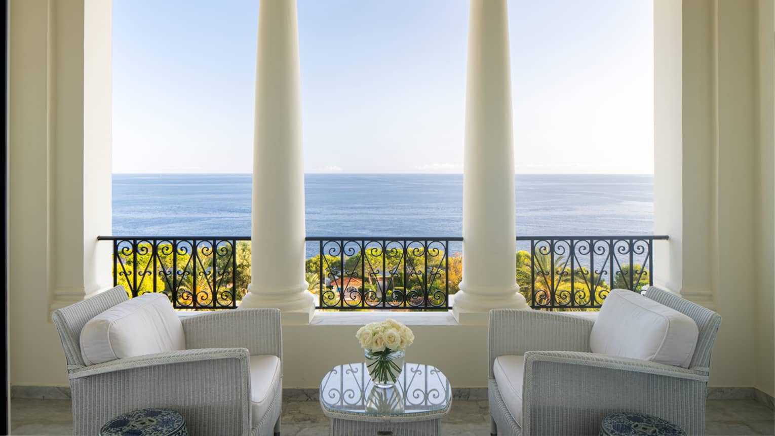 Two white club chairs facing with table between and beside white columns and terrace with sea view