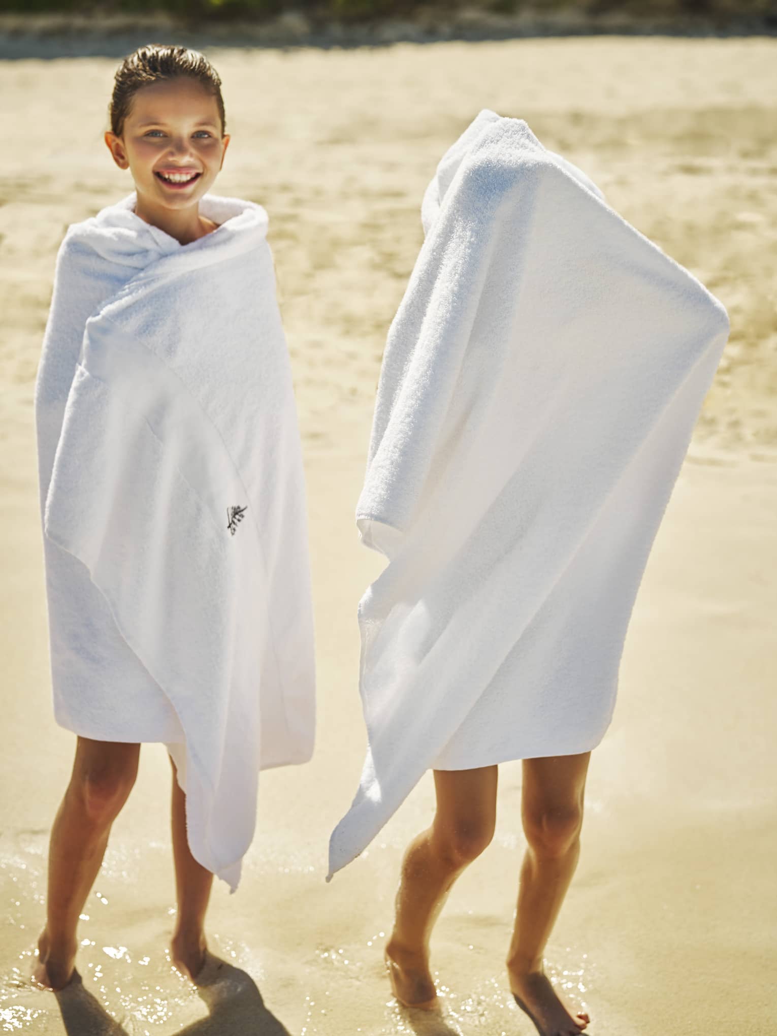 Two children standing on the beach with beach towels.
