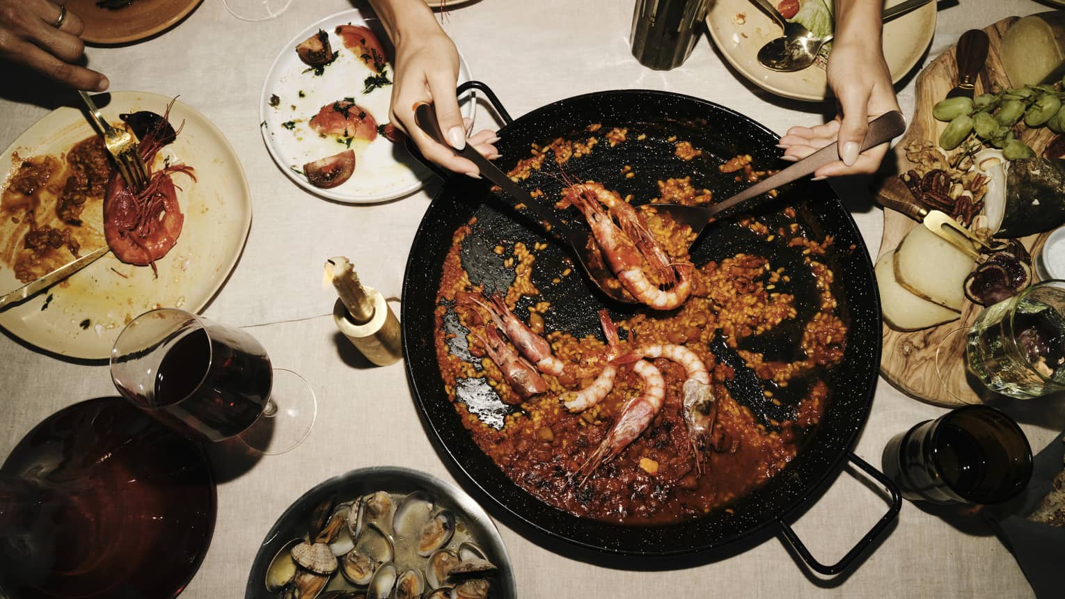 Two hands use large spoons to lift prawns from a saucy paella dish set amid plates of seafood, cheese and fruit.