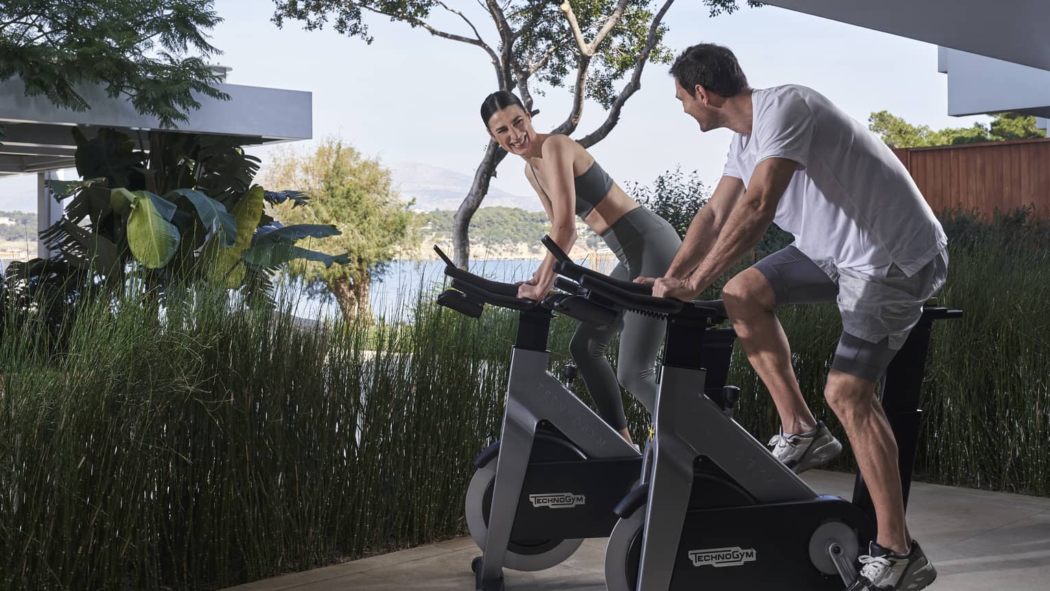 Couple riding stationary bikes on outdoor platform with water view in backdrop