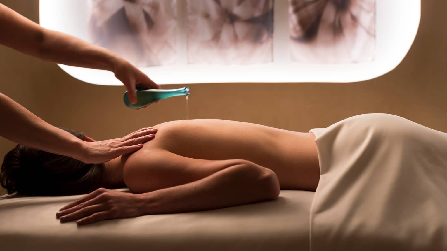 Woman's hands holding blue bottle pours liquid on woman's bare back in spa room