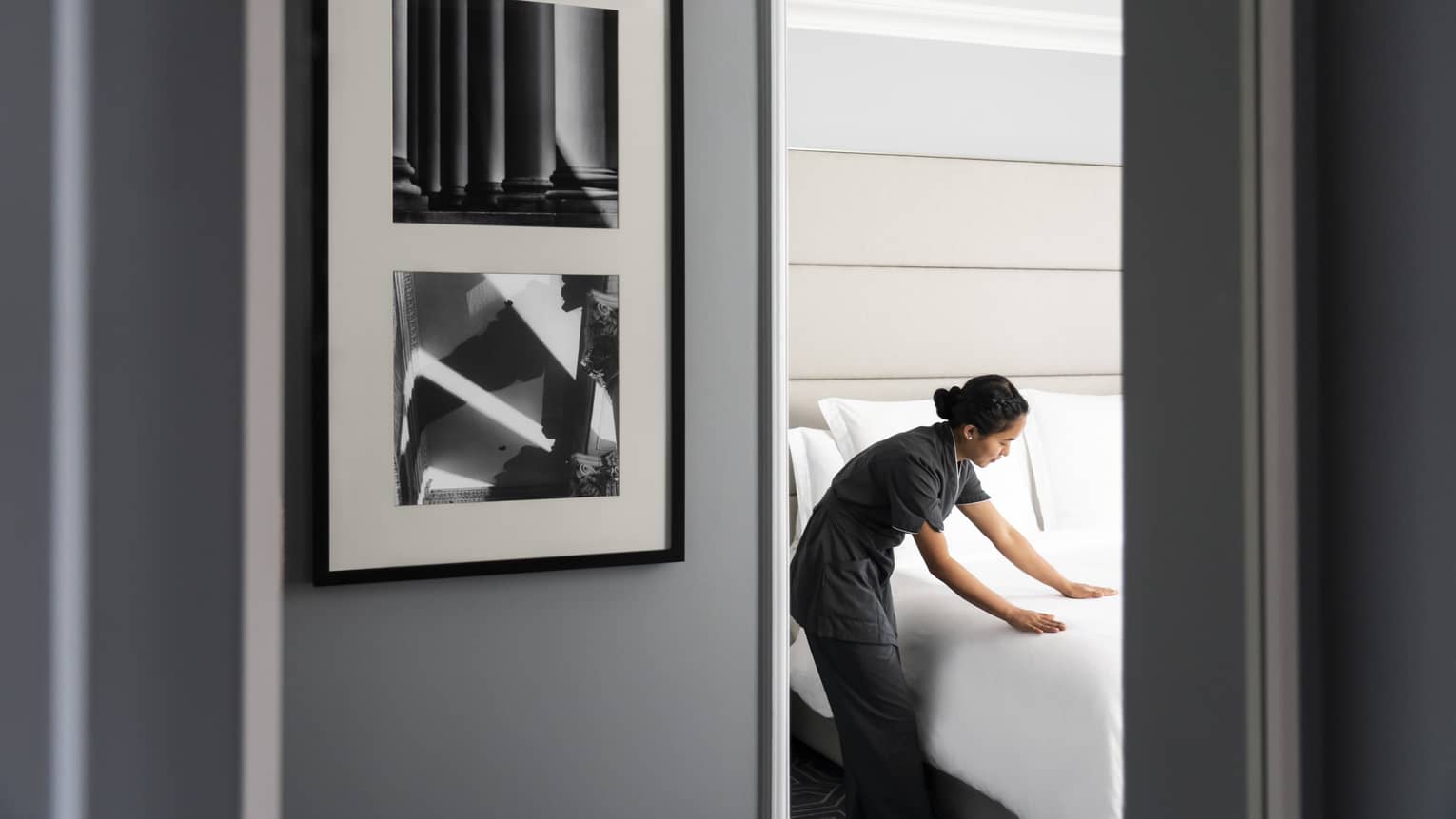 View past door and wall with framed art to housekeeping staff smoothing bed sheets